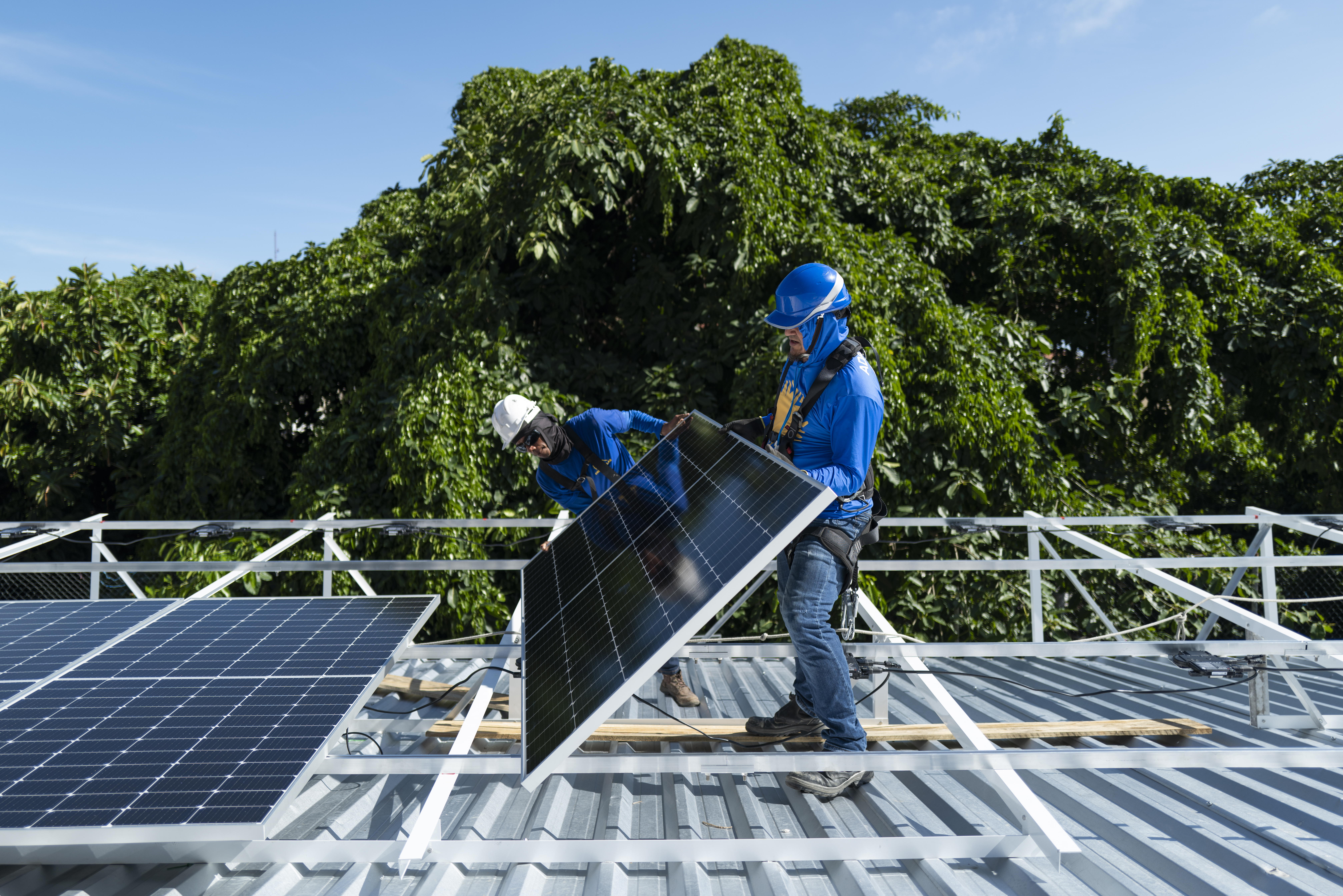 Två arbetare som installerar solpaneler på ett metalltak med träd i bakgrunden.