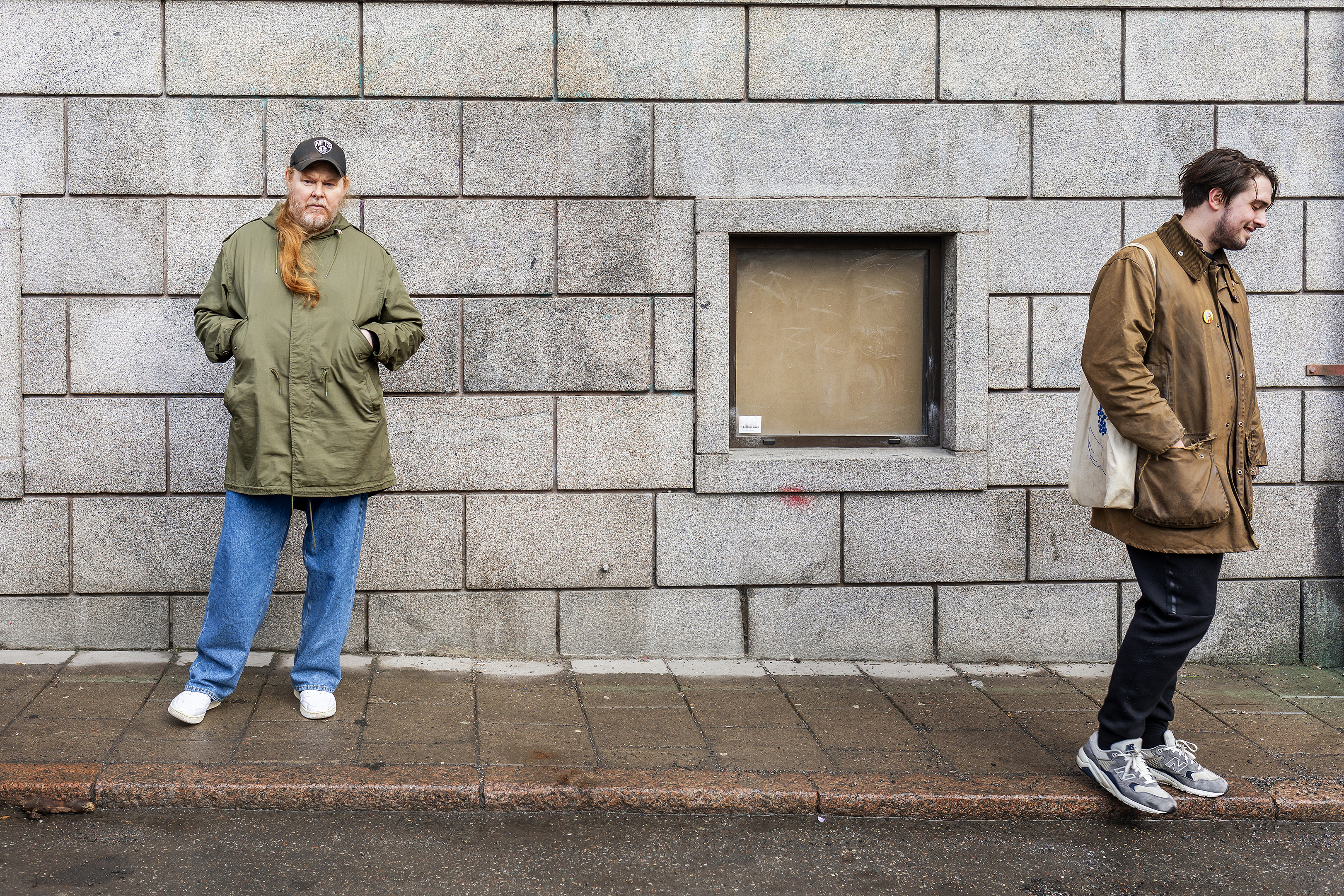 Två personer som går förbi varandra på en stadstrottoar med en grå kaklad vägg i bakgrunden.