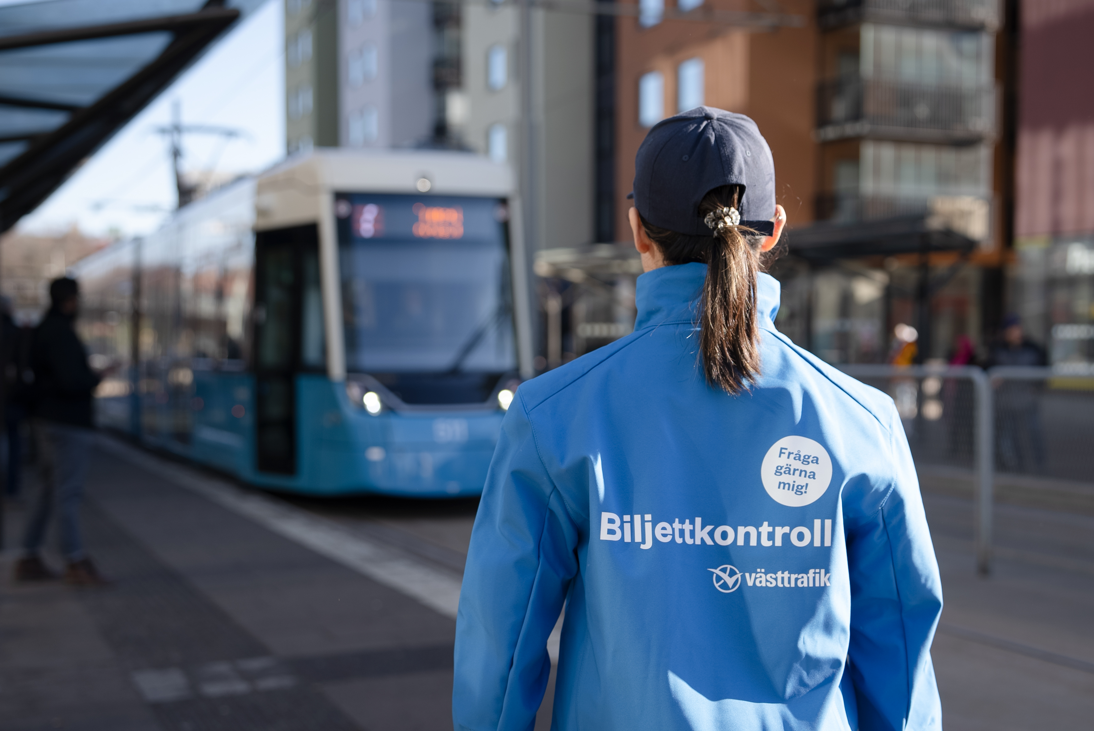 En biljettkontrollant i blå jacka med texten "biljettkontroll" står vänt mot en spårvagn på en stadsgata.