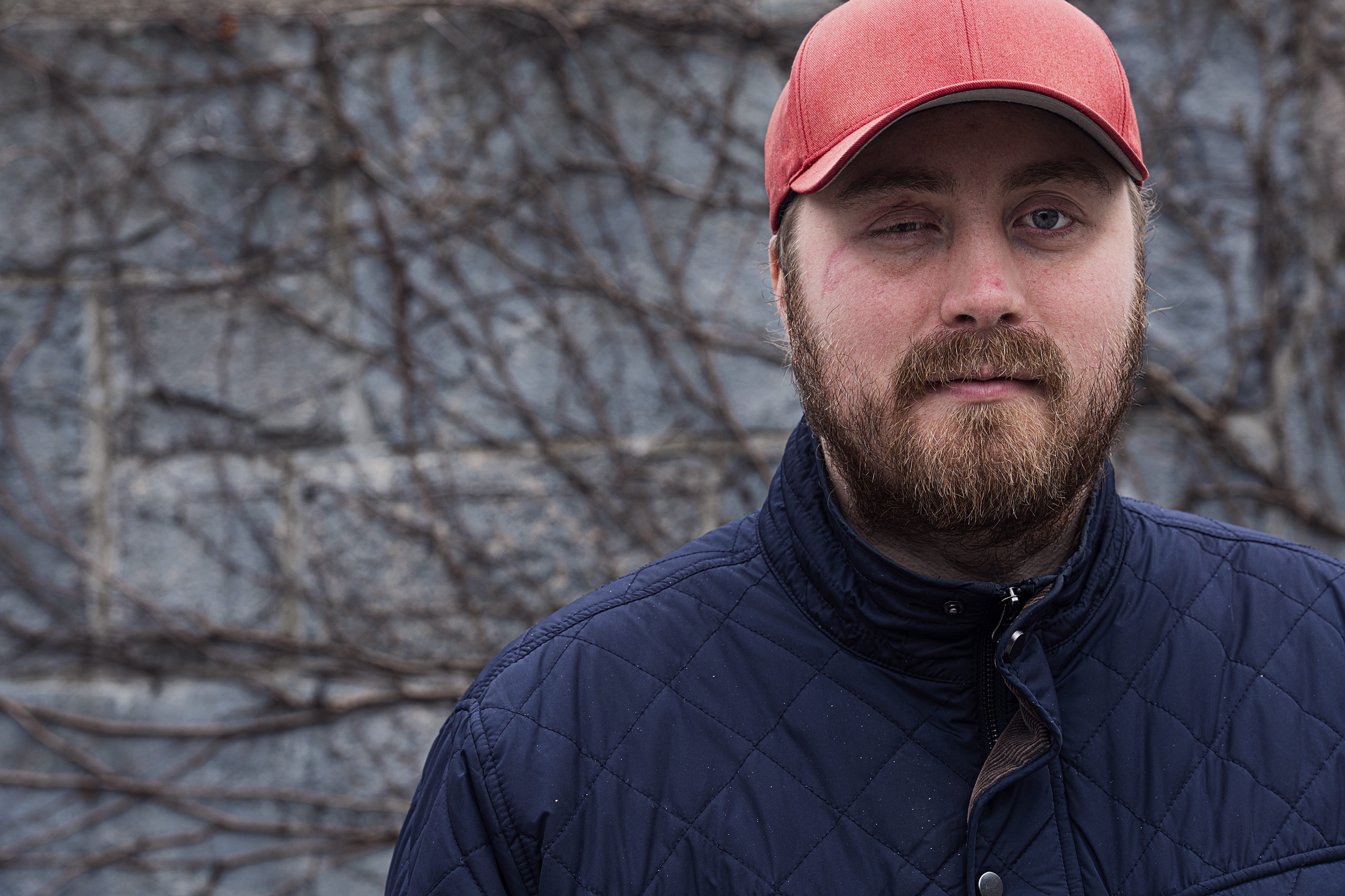 En skäggig man i röd keps och marinblå jacka som står med kala buskar i bakgrunden.