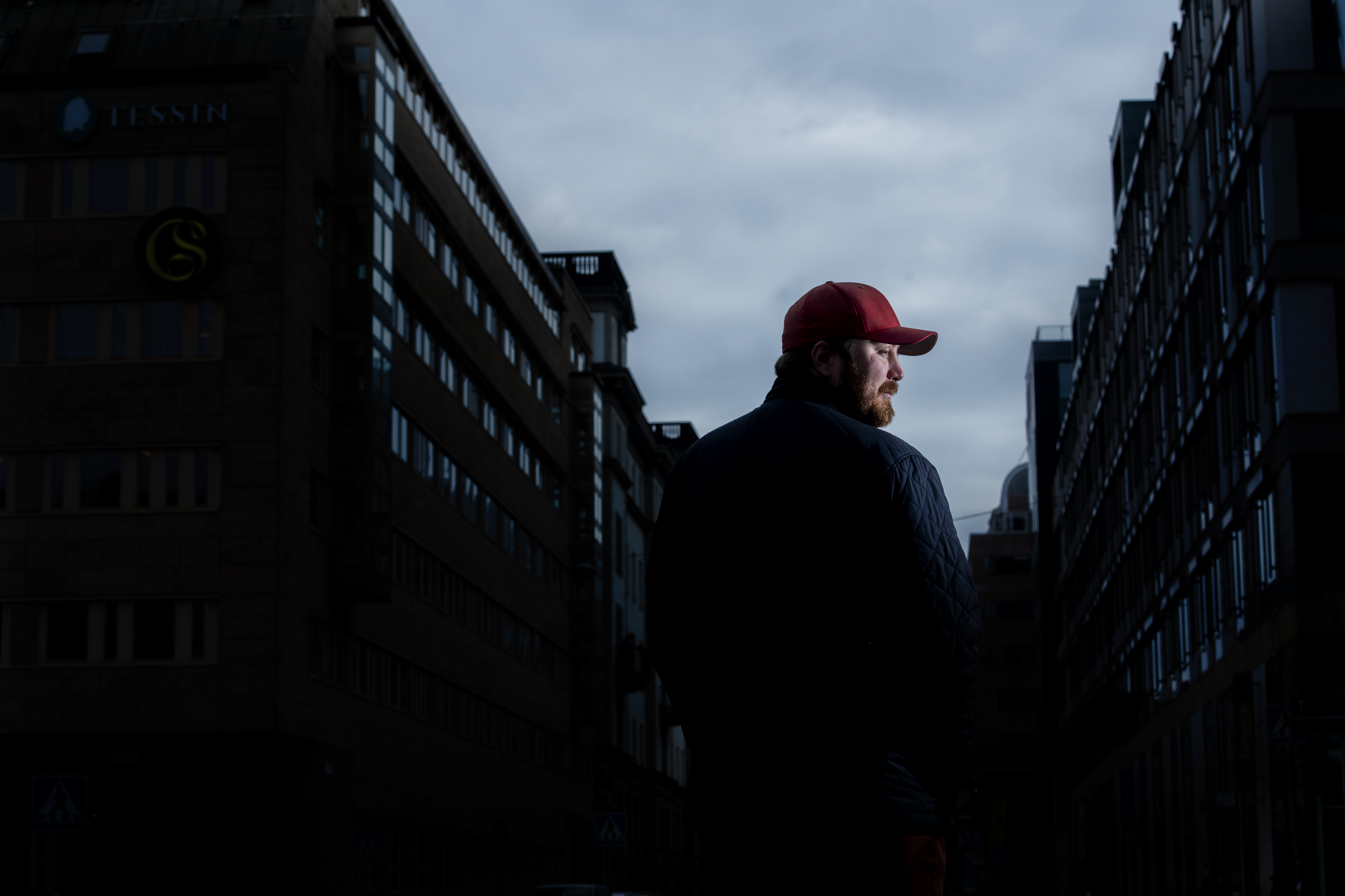 En skäggig man i en röd keps står i profil med urbana byggnader under ett molnigt himlaval som bakgrund.