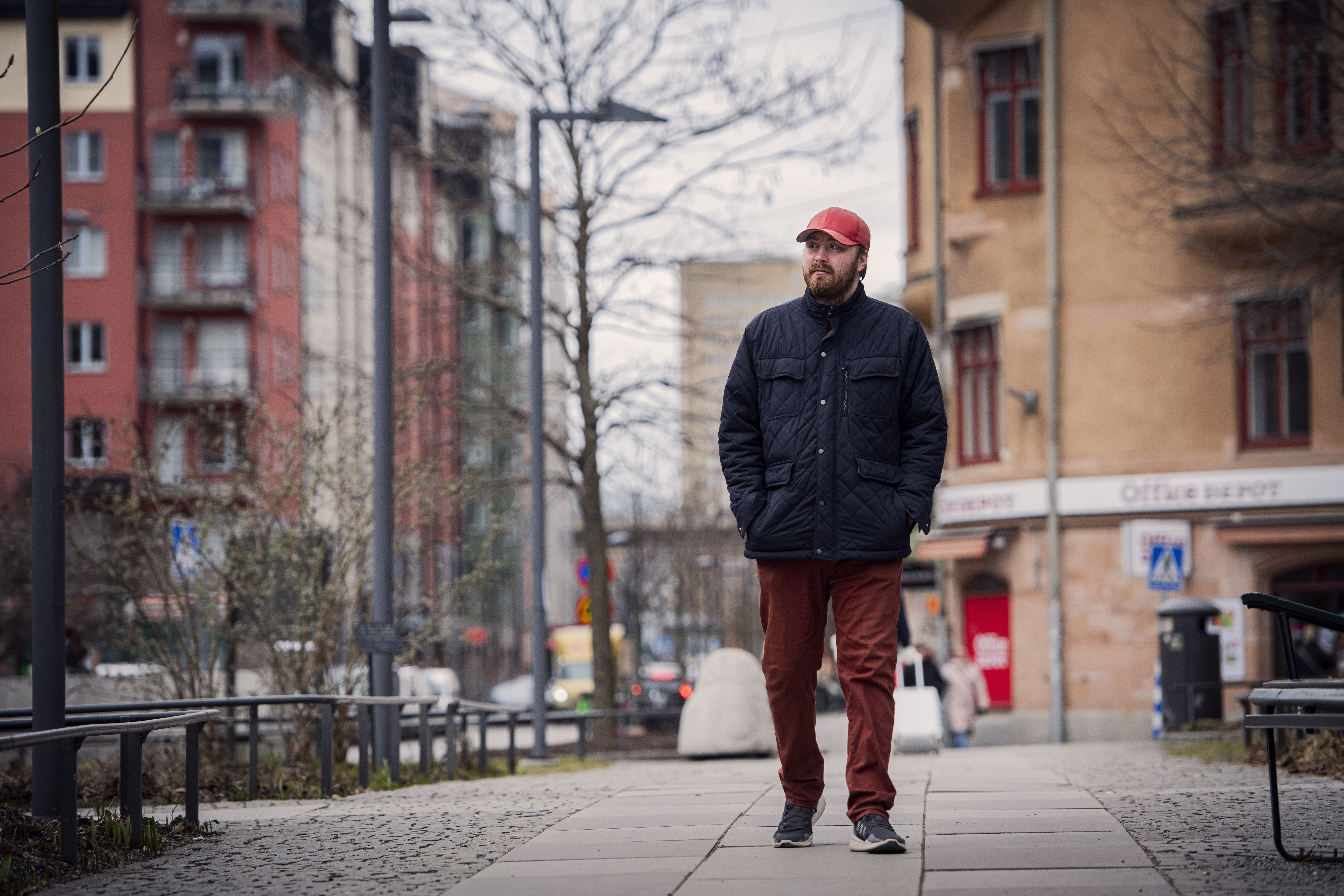 En man klädd i en röd keps, svart jacka och röda byxor promenerar på en trottoar i ett stadsområde med byggnader i bakgrunden.