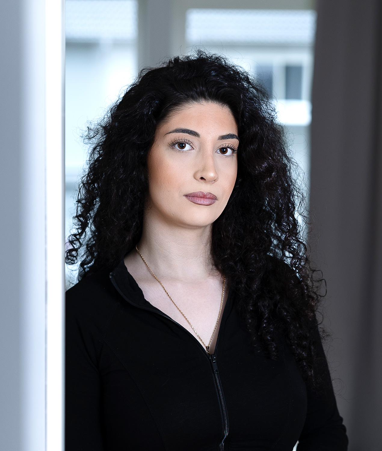 En kvinna med lockigt svart hår och svart topp står vid ett fönster, blickar rakt in i kameran.