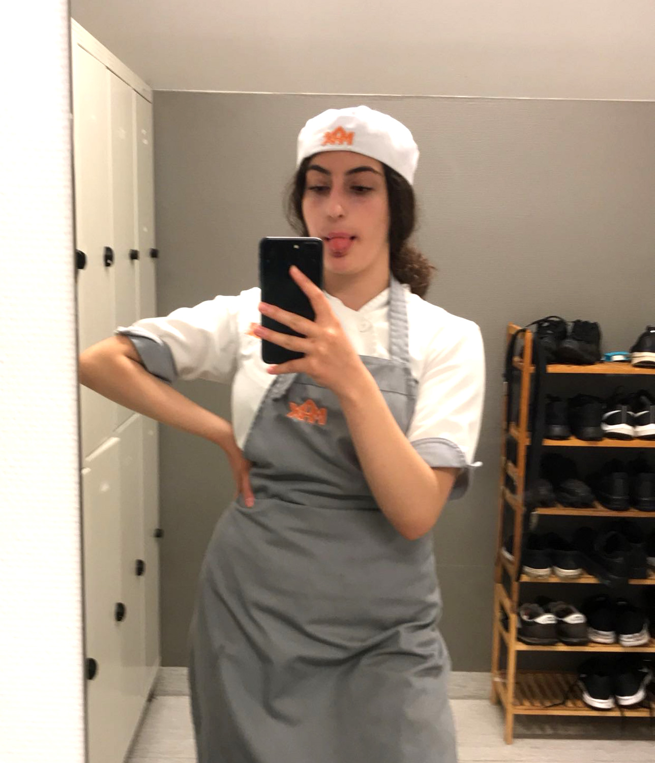 En person i Max-uniform inklusive en hatt med logotyp tar en spegelselfie medan de poserar med en hand på höften i ett omklädningsrum.