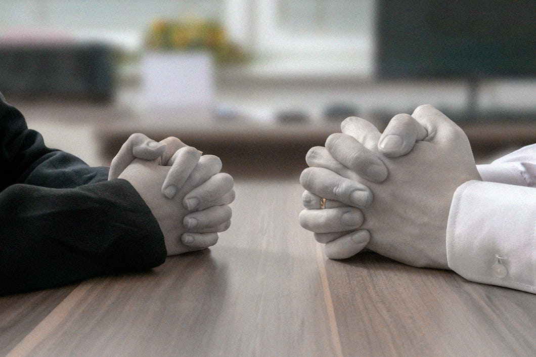 Två individer som sitter mittemot varandra, med händerna sammanslagna på ett bord, i en gest som antyder en seriös konversation eller förhandling.