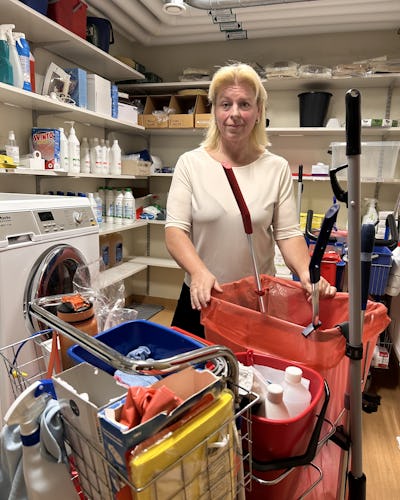 Kvinna sorterar städmaterial i ett förråd med hyllor och olika städutrustning.
