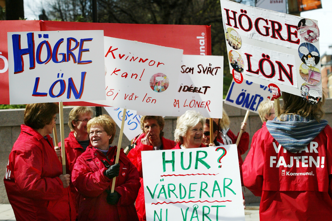 Demonstranter i röda jackor håller skyltar som förespråkar högre löner och jämställdhet i löner.