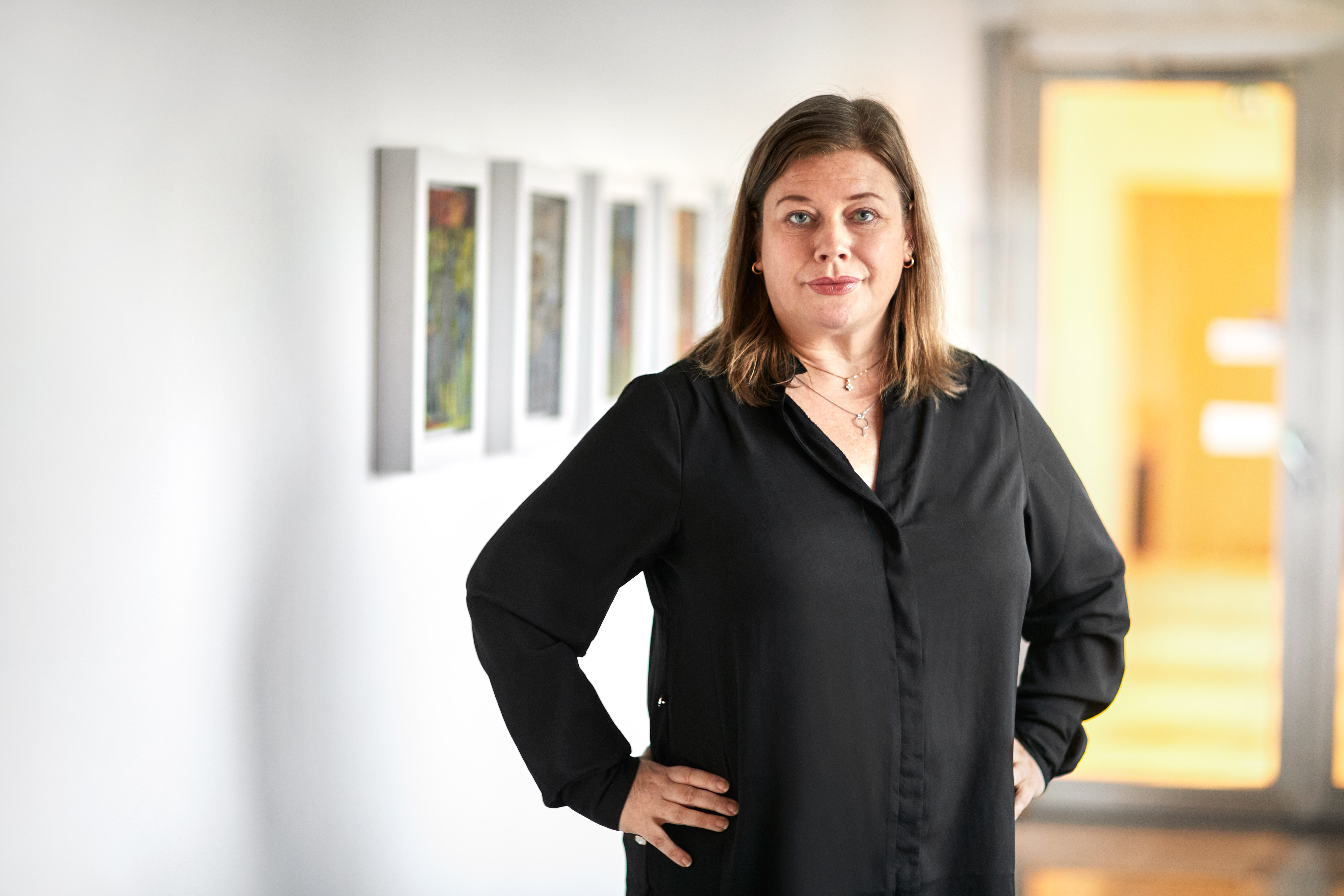 Malin Ragnegård, Kommunals ordförande, i svart blus står i ett konstgalleri med händerna på höfterna, med inramade bilder som hänger på väggen bakom henne.