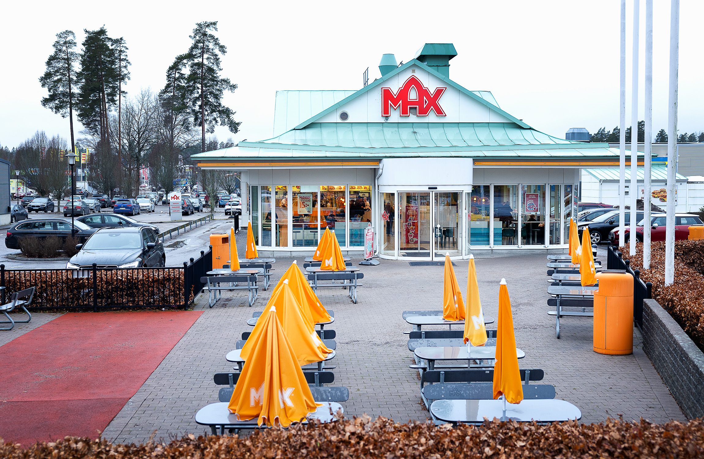 Alt text: Max snabbmatsrestaurang exteriör med uteservering, orange paraplyer, i en förortsmiljö med parkerade bilar och träd synliga i bakgrunden.