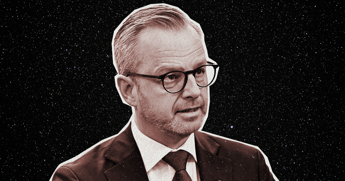 En man med blont hår och glasögon klädd i en mörk kostym. Bakom är en mörk himmel med stjärnor.