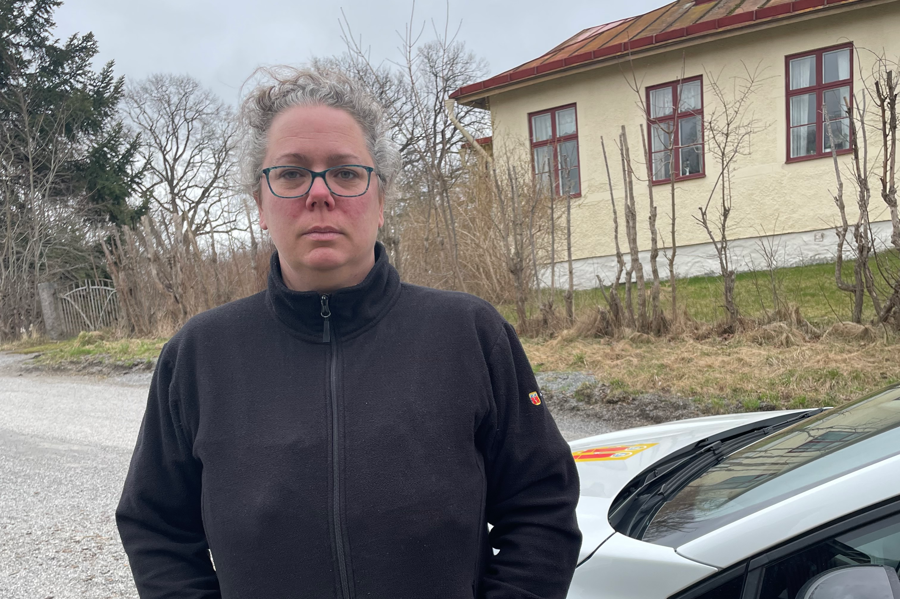 Medelålders kvinna med lockigt grått hår och glasögon står framför ett hus med rött tak, delvis skymd av en bil.