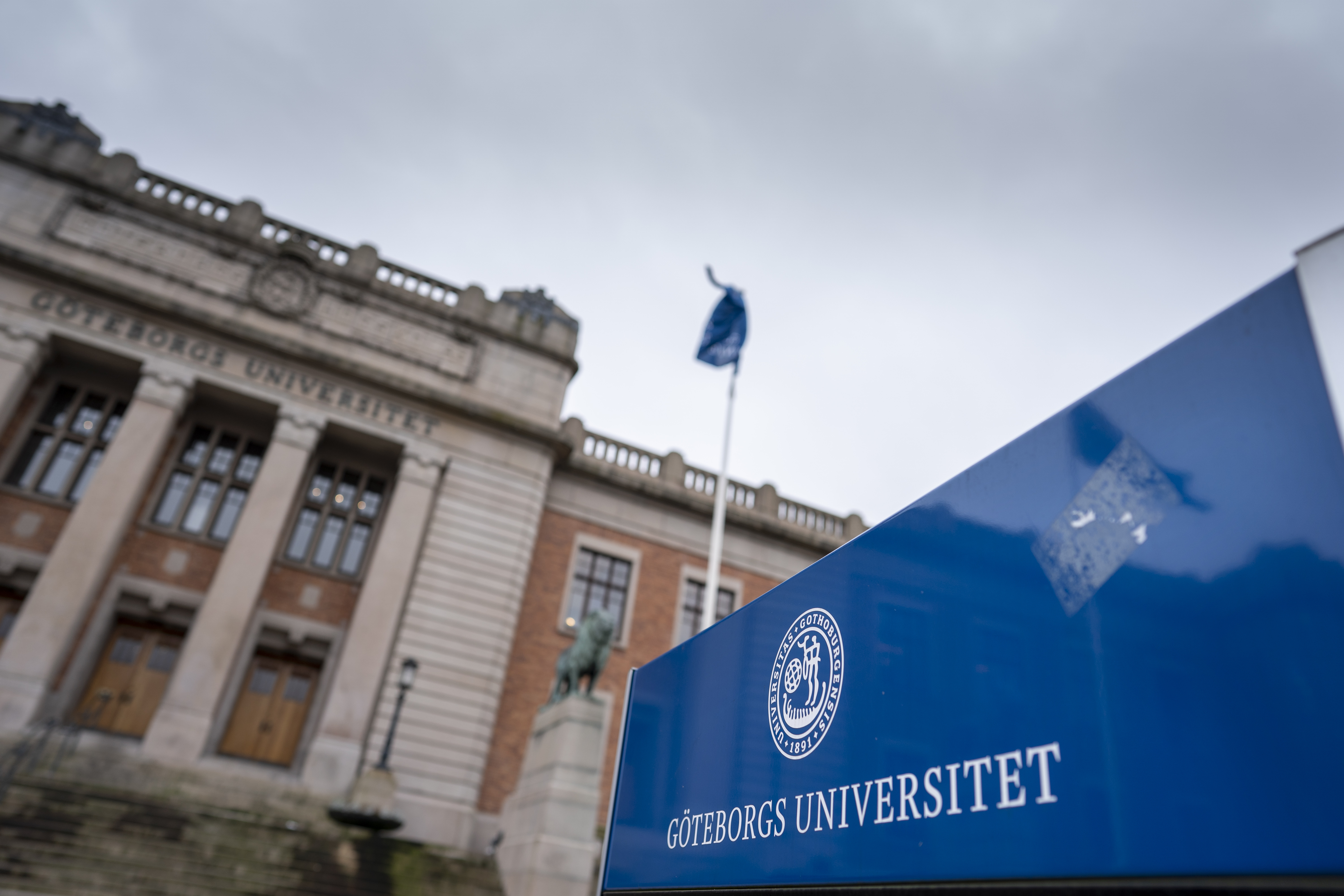 Skylt som visar Göteborgs universitets logotyp i förgrunden, med den historiska universitetsbyggnaden något oskarp i bakgrunden.