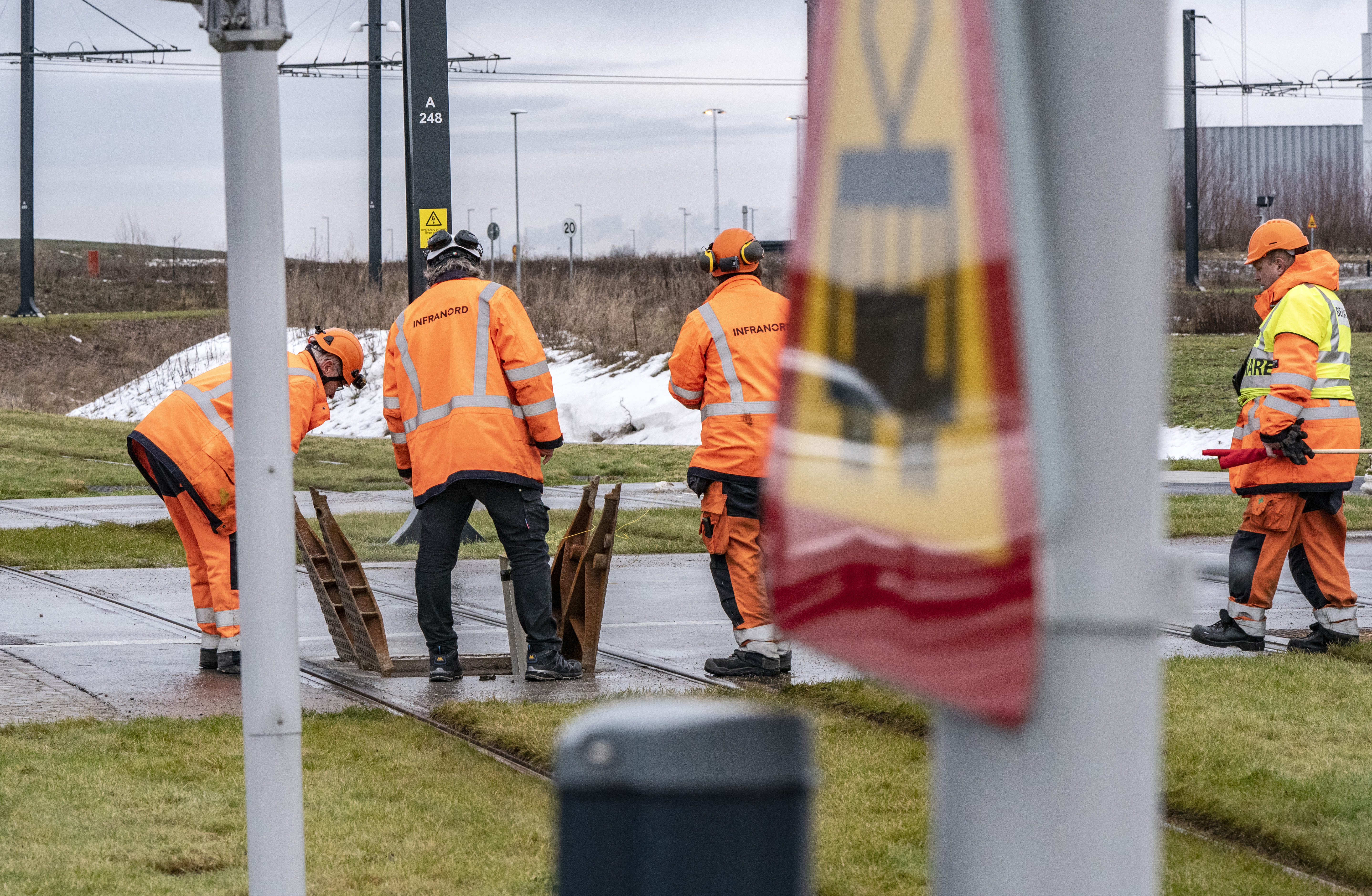 Fyra banarbetare med orange Infranord-jackor arbetar vid ett järnvägsspår, med en tågskylt i förgrunden.