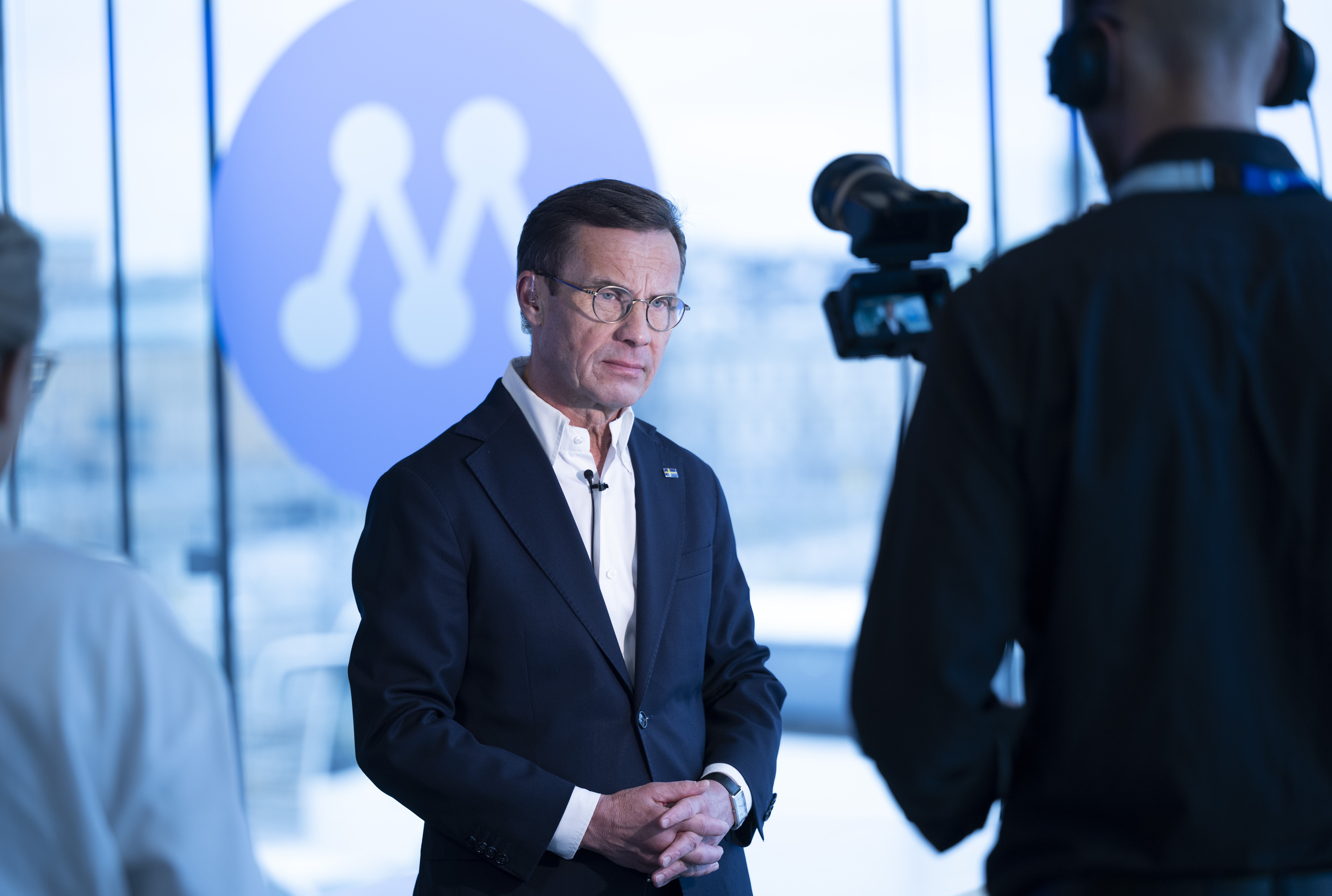 Person i kostym intervjuas på kameran, står framför ett fönster med en logotyp i bakgrunden.