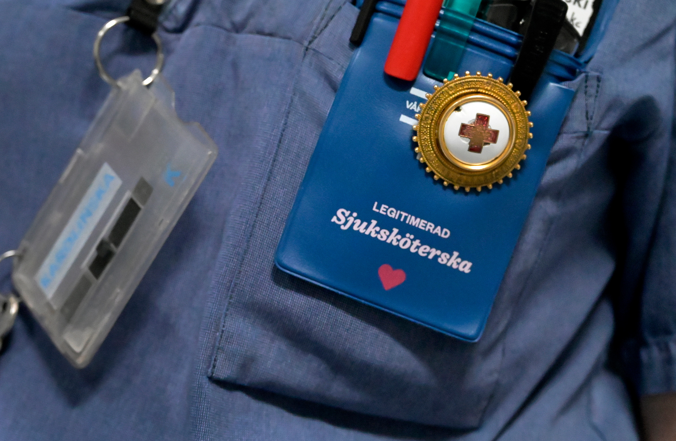En närbild på en sjuksköterskas märke och pennor i en ficka, där identifikation och en röd hjärtsymbol syns, fäst på en blå uniform.
