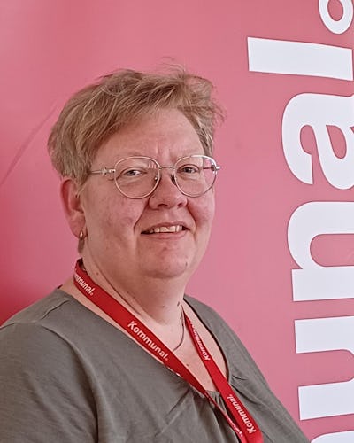 Anette Modin med kort hår, glasögon och en röd nyckelrem står framför en röd bakgrund med delvis synlig vit text.