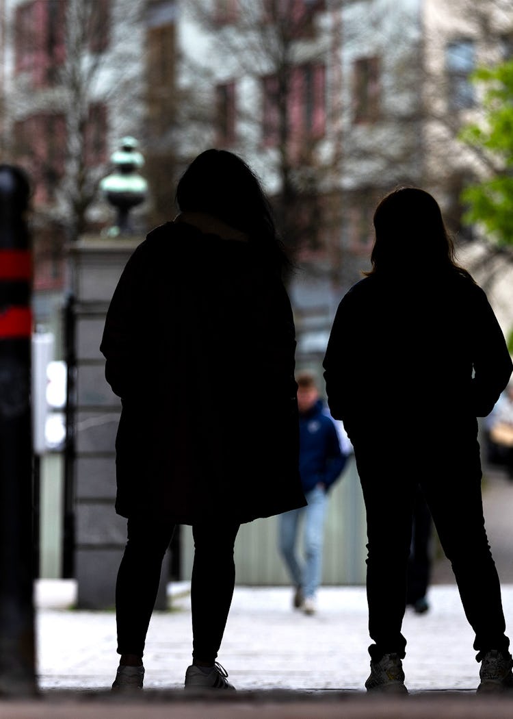 Två personer står i en skuggad gränd med en stadsgata i bakgrunden. Andra fotgängare kan ses gå längre fram.