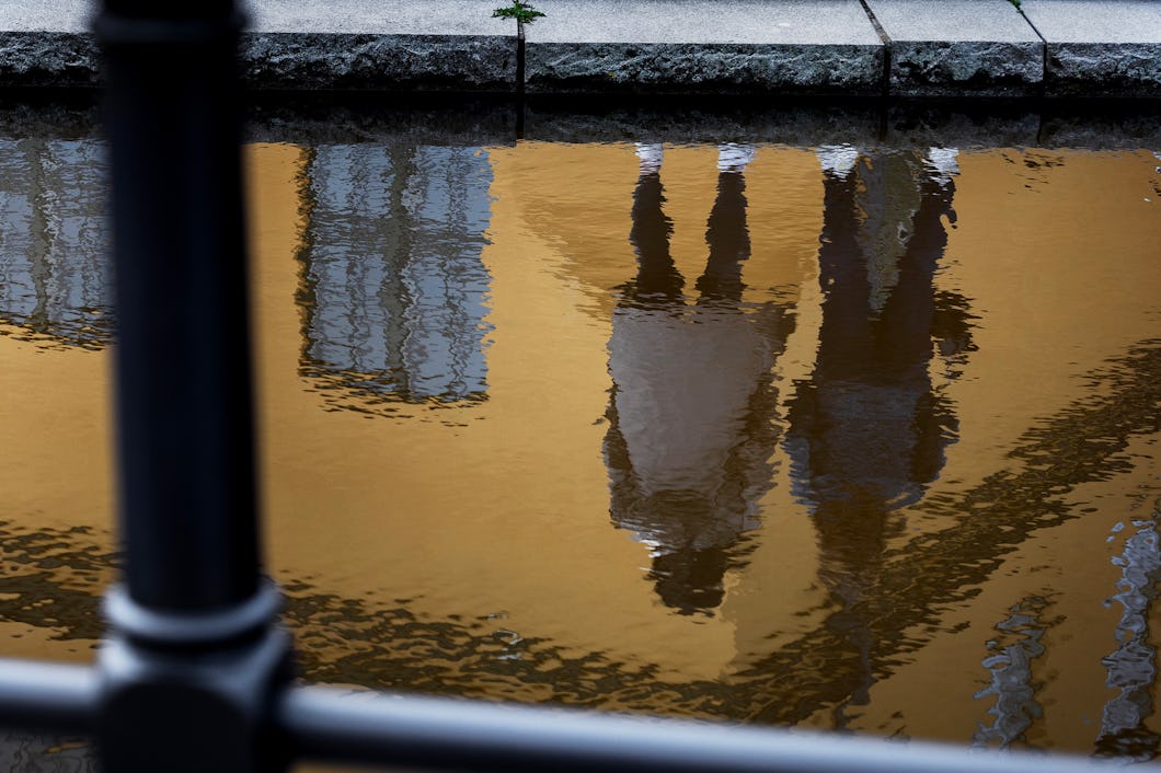 Två personer står nära en kanal, reflekterade i vattnet. I bakgrunden syns byggnader och trottoaren ovanför.