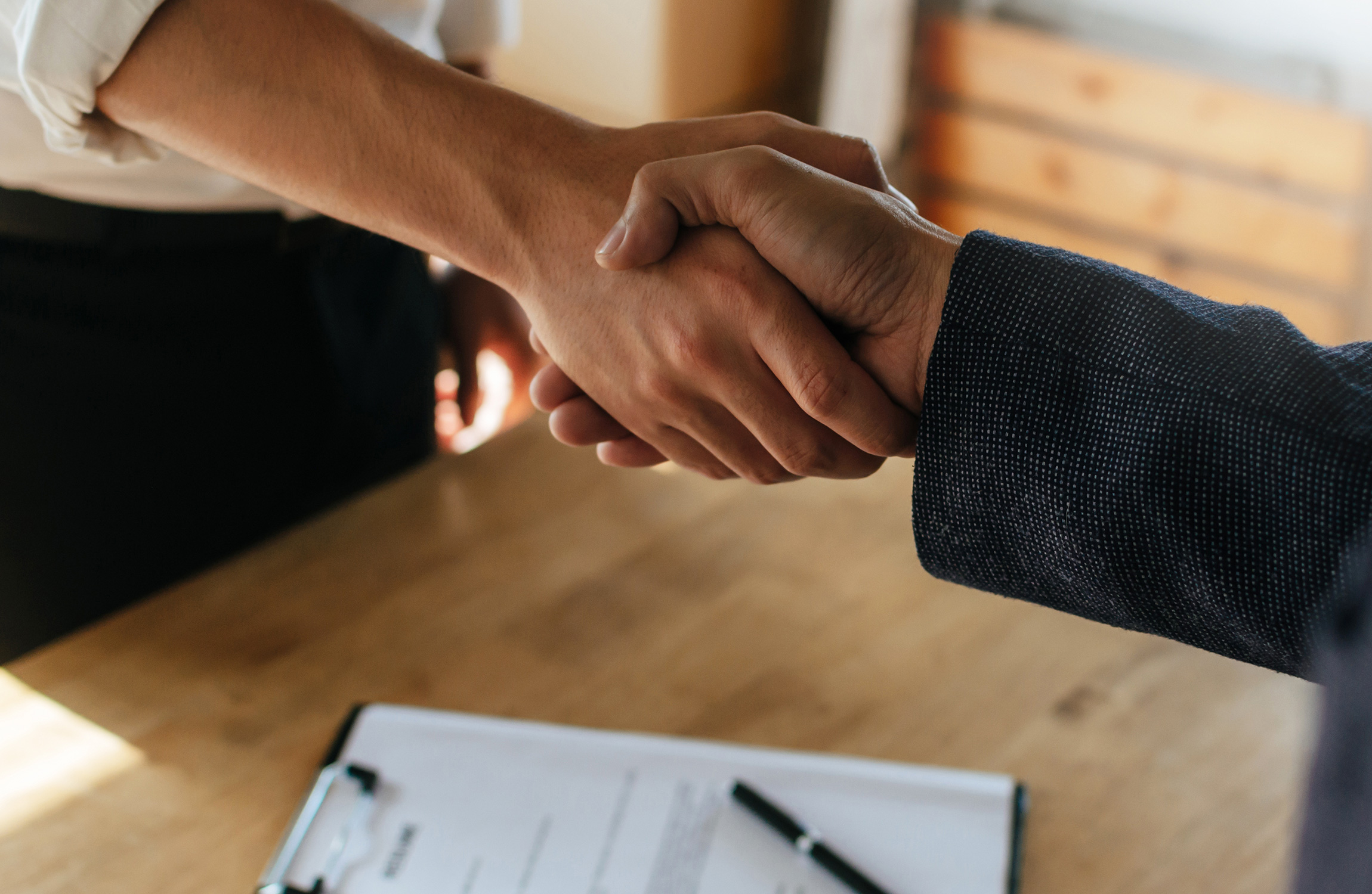 Två personer skakar hand över ett träbord med en clipboard och penna på.