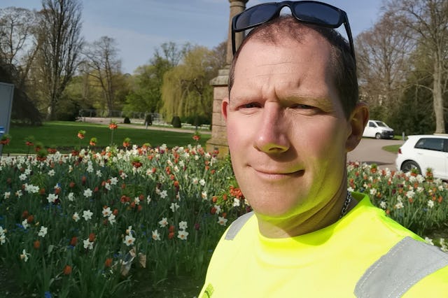 En man med solglasögon och en neongul skjorta håller sin smartphone för en selfie i en park. Scenen inkluderar blommande påskliljor och en staty i bakgrunden.