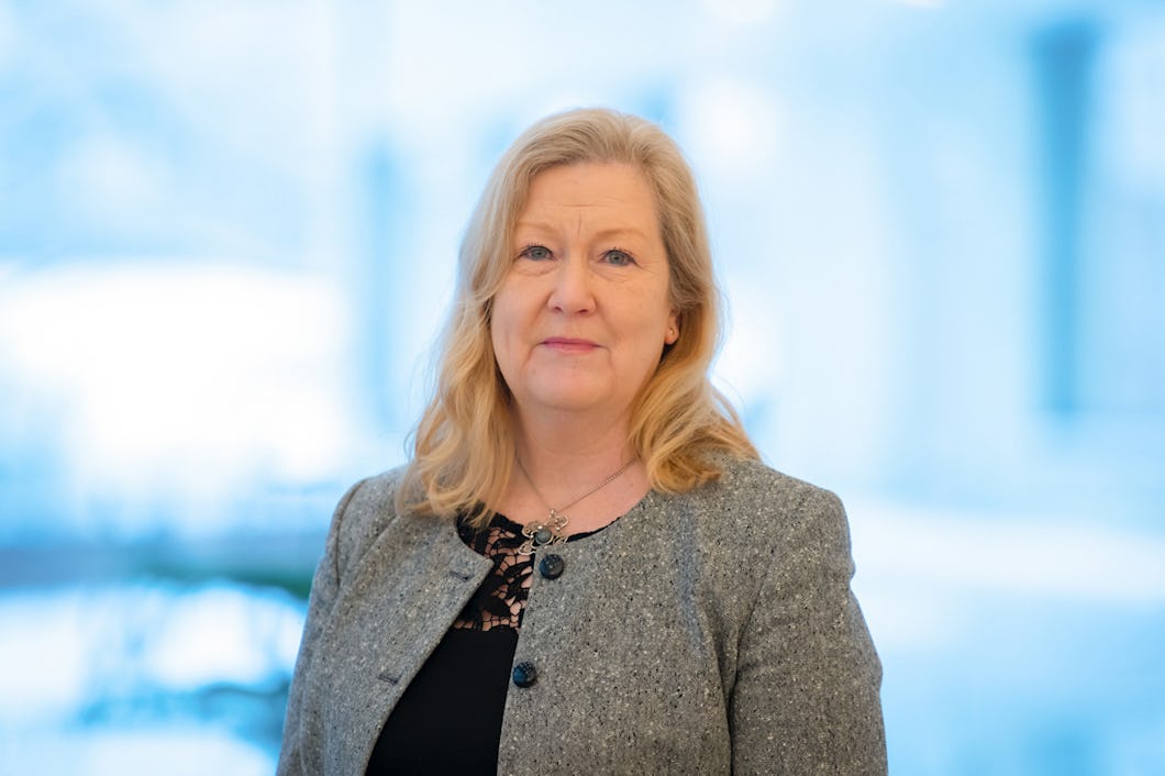 En kvinna med blont hår som har på sig en grå kavaj och en svart topp står mot en suddig blå bakgrund.