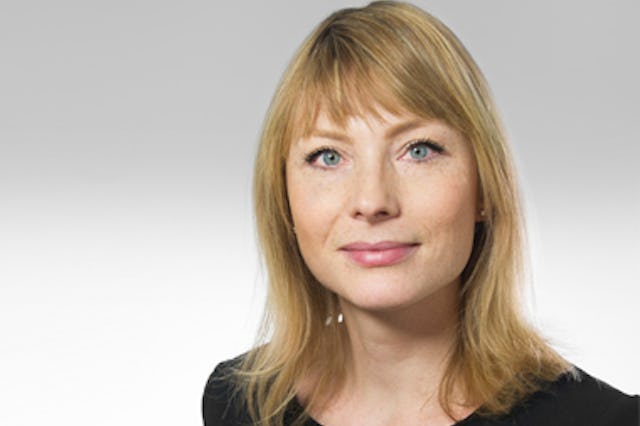 En professionell porträttbild av en kvinna med blont hår och lugg, blå ögon, bär diskret smink och små örhängen på en ljus bakgrund.