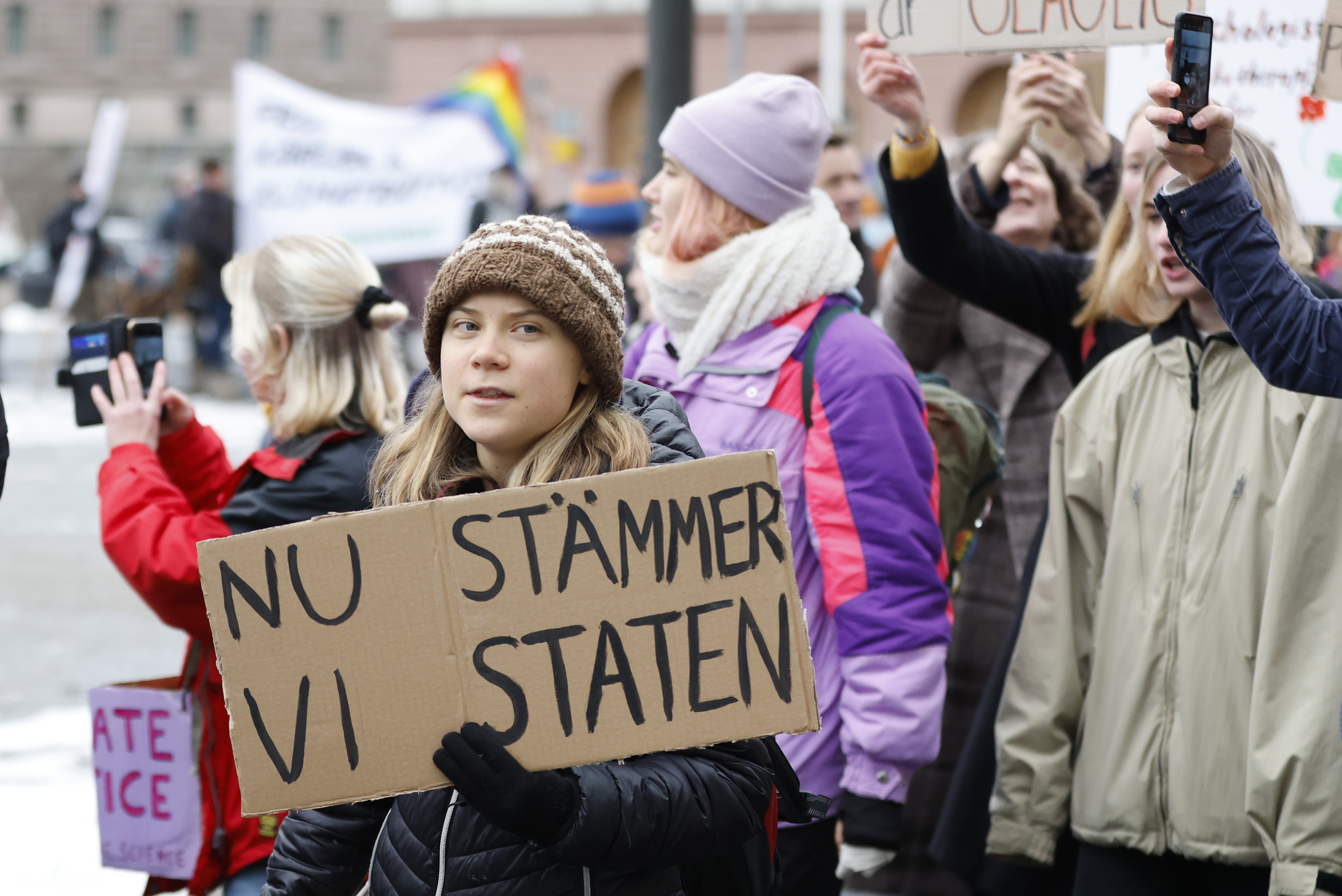 Alt text: En ung kvinna (Greta Thunberg) håller ett protestplakat med texten "nu stämmer vi staten" på en offentlig demonstration, omgiven av andra demonstranter med plakat och kameror.