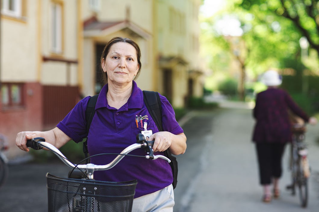 Fadime Mavi i lila skjorta, med en ryggsäck och en cykel, står på en gata med byggnader och träd i bakgrunden. En annan individ går bort med en cykel.