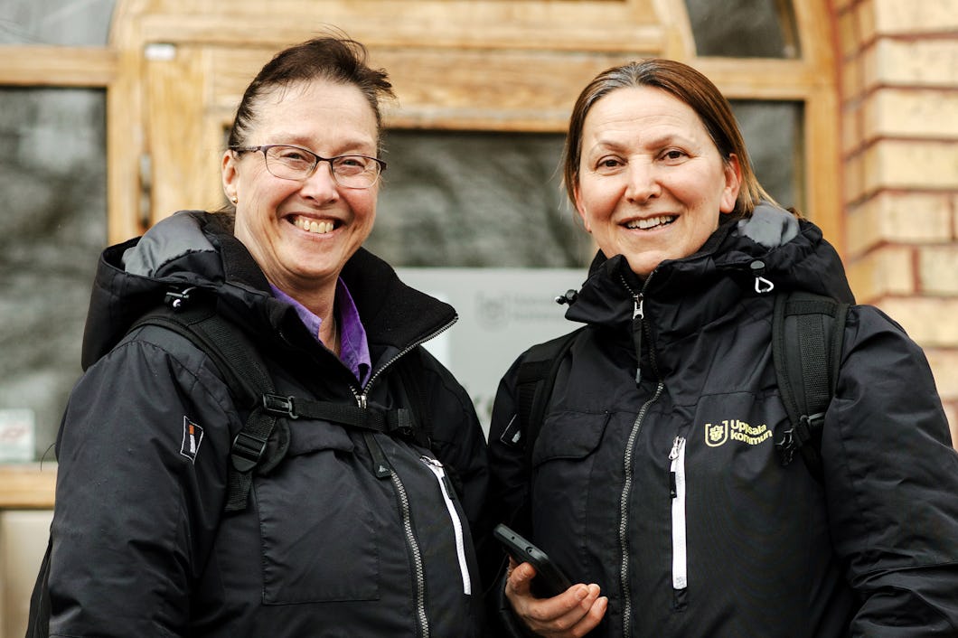 Två kvinnor i svarta jackor och ryggsäckar står framför en träbyggnad och ler mot kameran.