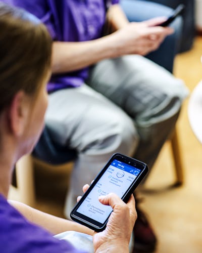 Två personer använder smartphones i ett väntrum, med fokus på en kvinnas händer som betraktar en hälsorelaterad app på sin enhet.