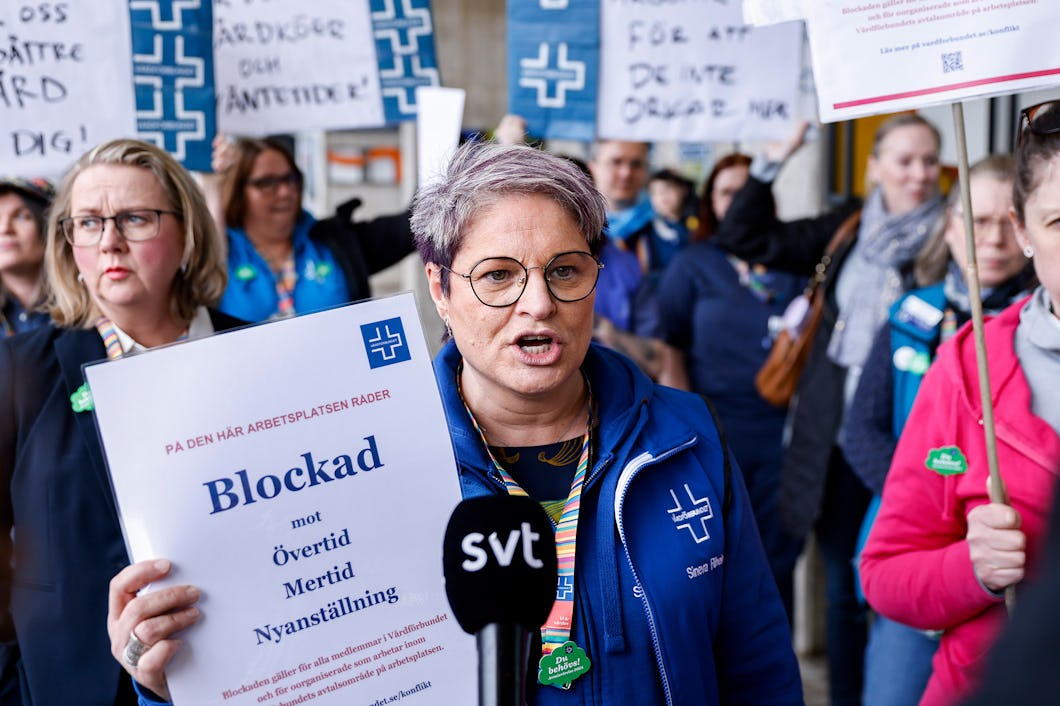 En grupp håller människor upp protestskyltar. En person med kort, lila hår talar i en mikrofon märkt "svt".