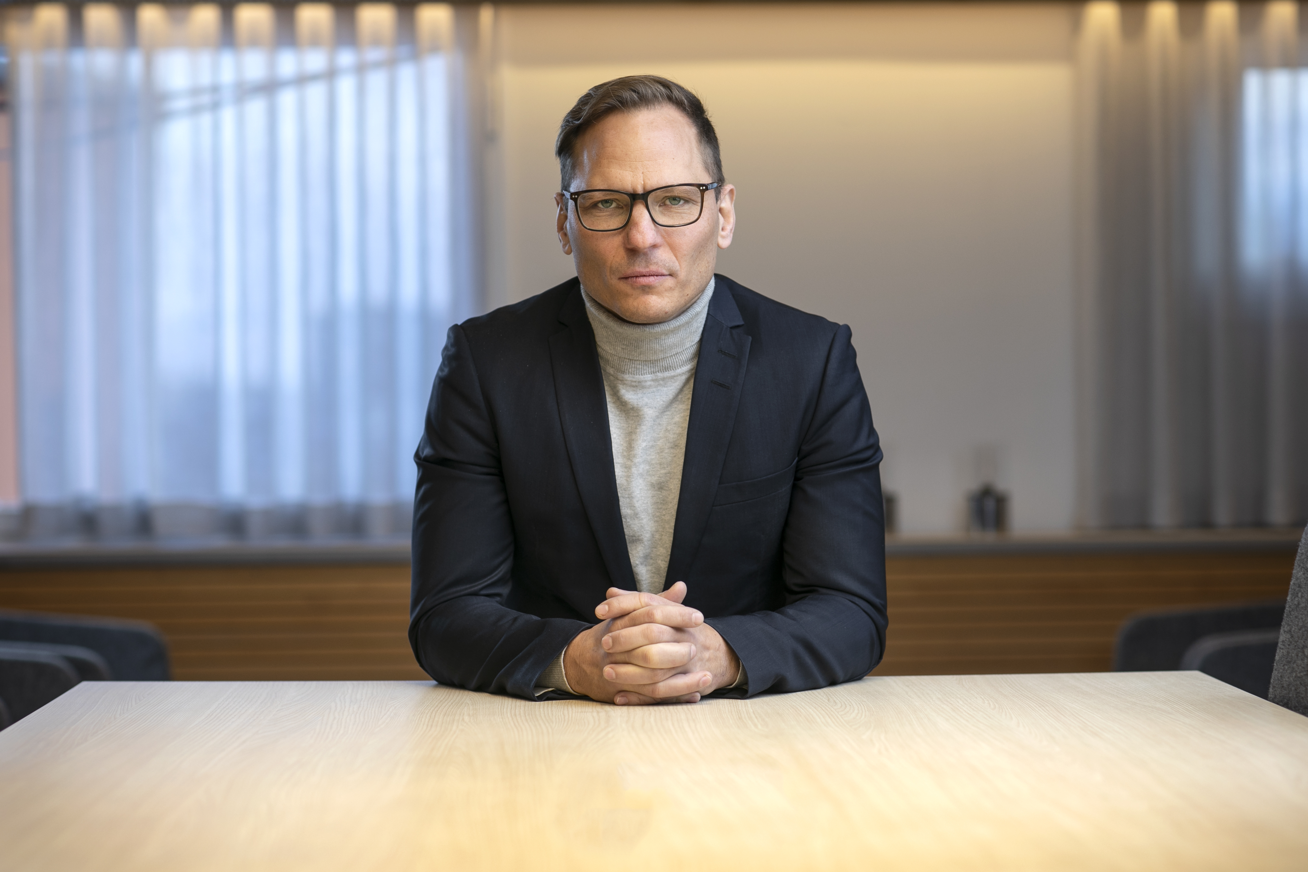 Johan Ingelskog med glasögon, klädd i en svart kavaj och grå polotröja, sitter vid ett bord med händerna knäppta, vänd mot kameran. Rummet har genomskinliga gardiner och belysta väggdetaljer i bakgrunden.