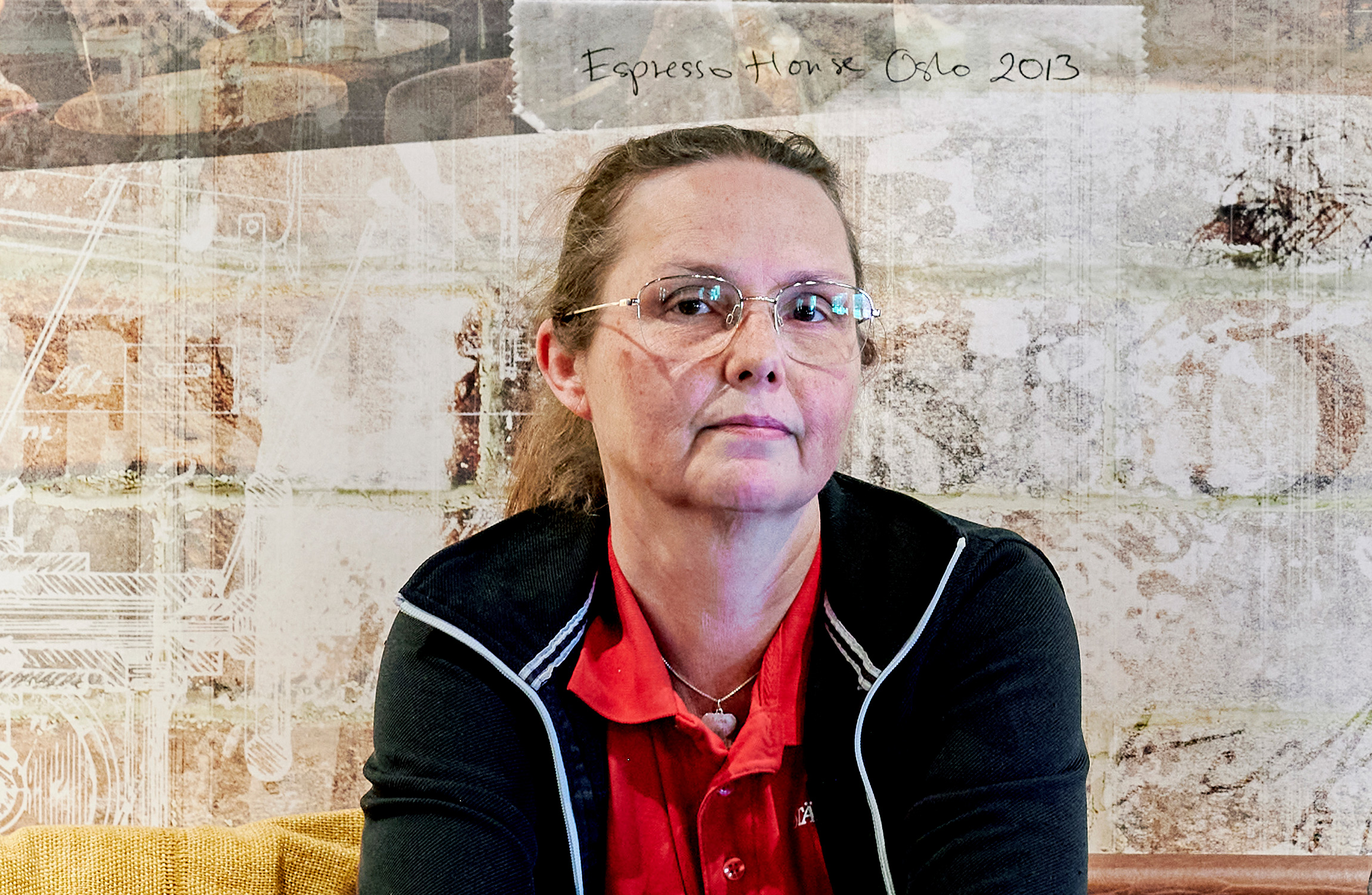 En person med glasögon och en röd skjorta sitter mot en vägg med texten "Espresso Hon x Oslo 2013" i bakgrunden.