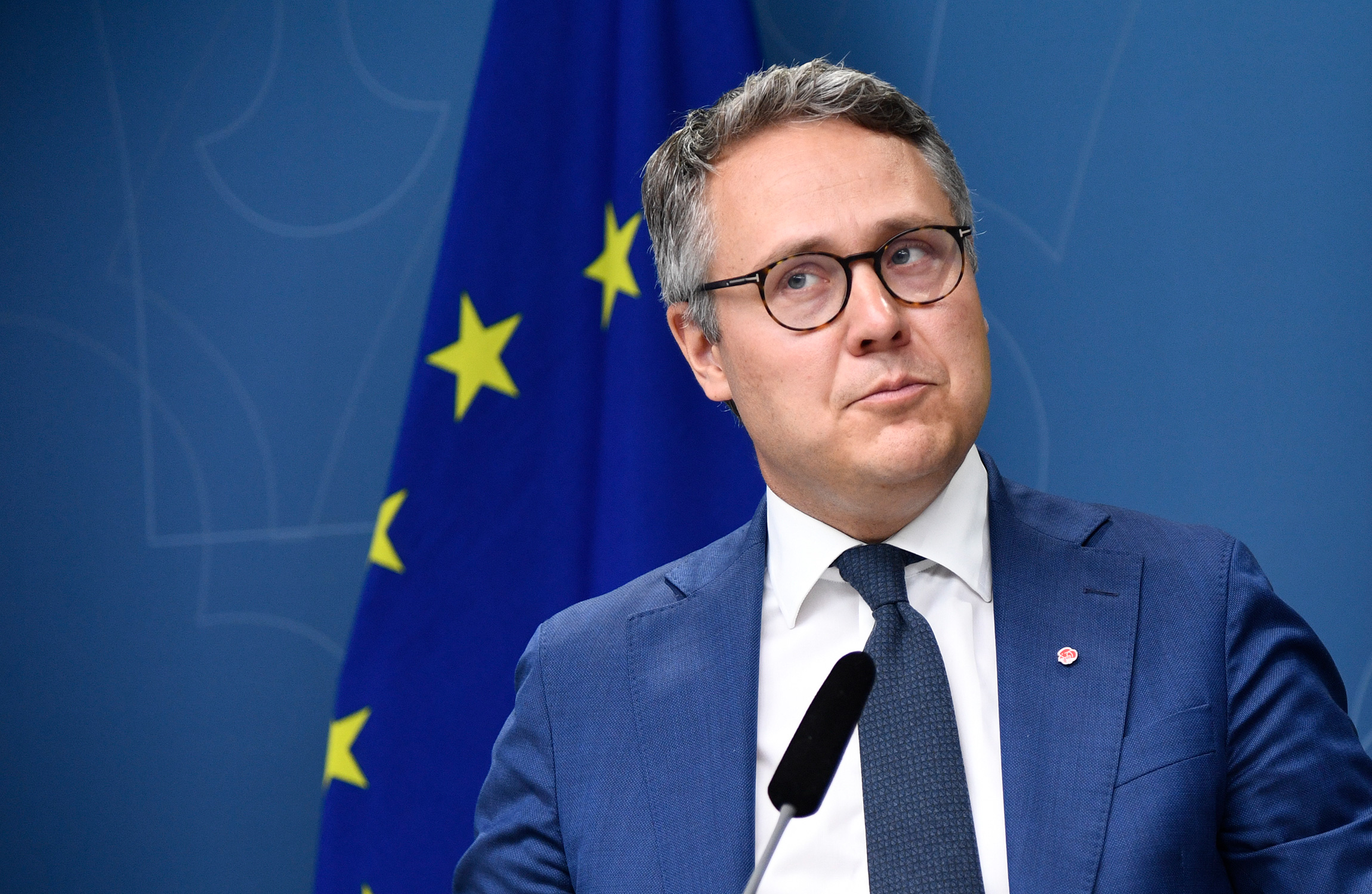 En man i kostym och slips står framför en EU-flagga. Han talar vid ett podium med en mikrofon.