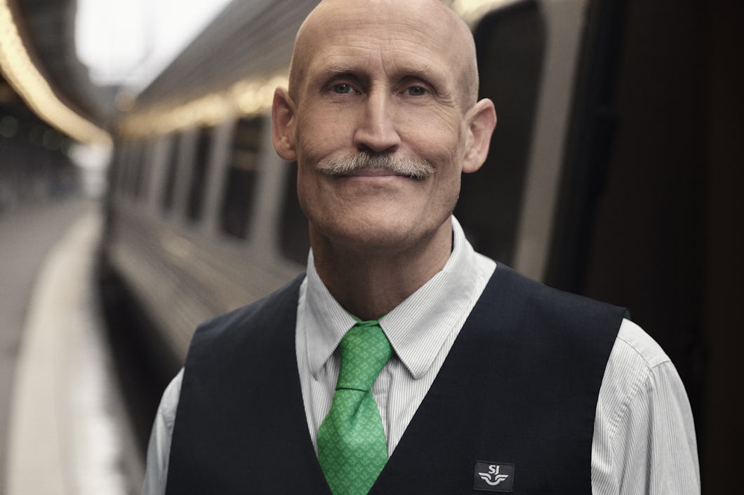Lars Fuhre iklädd en vit skjorta, grön slips och svart väst med "SJ"-logga, står framför ett tåg på en perrong.