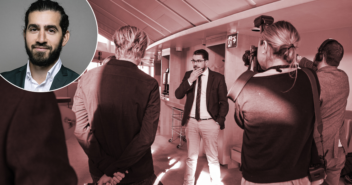 En man i kostym talar framför ett kamerateam och några åskådare i en inomhusmiljö. I det övre vänstra hörnet finns en liten porträttbild av en annan man med skägg.