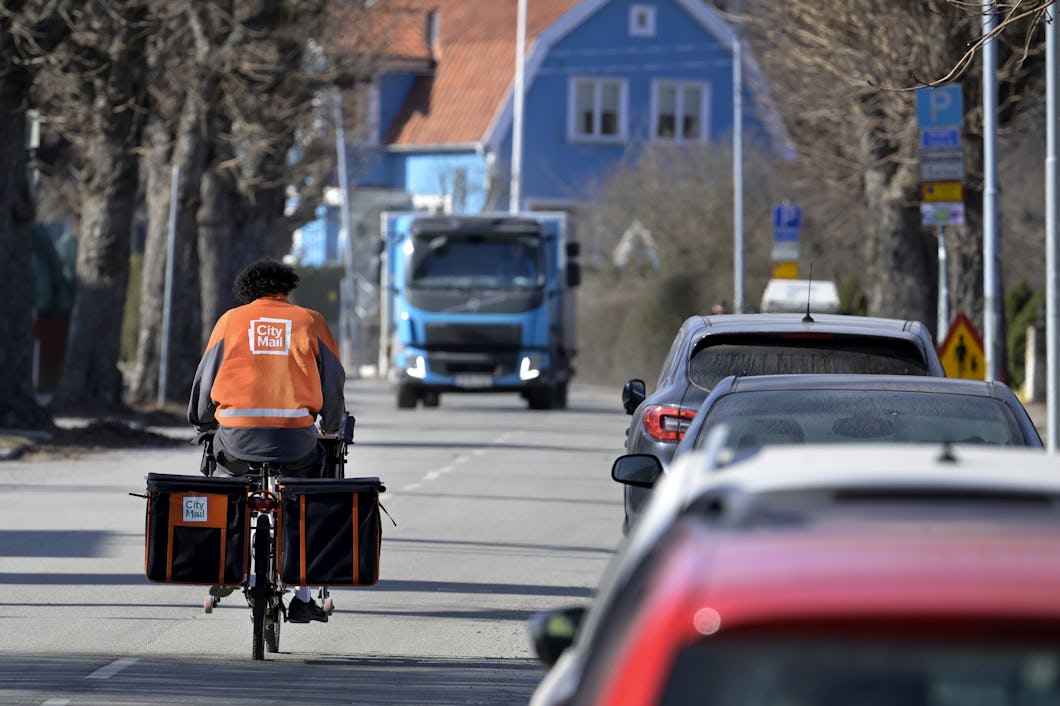 En brevbärare på cykel från Citymail cyklar längs en gata och är på väg att möta en blå lastbil från Postnord.