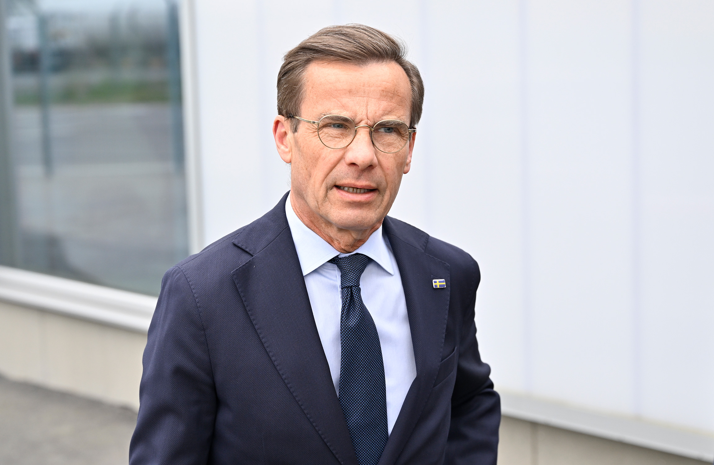 En man i kostym och slips, med glasögon, står utomhus framför en modern byggnad.