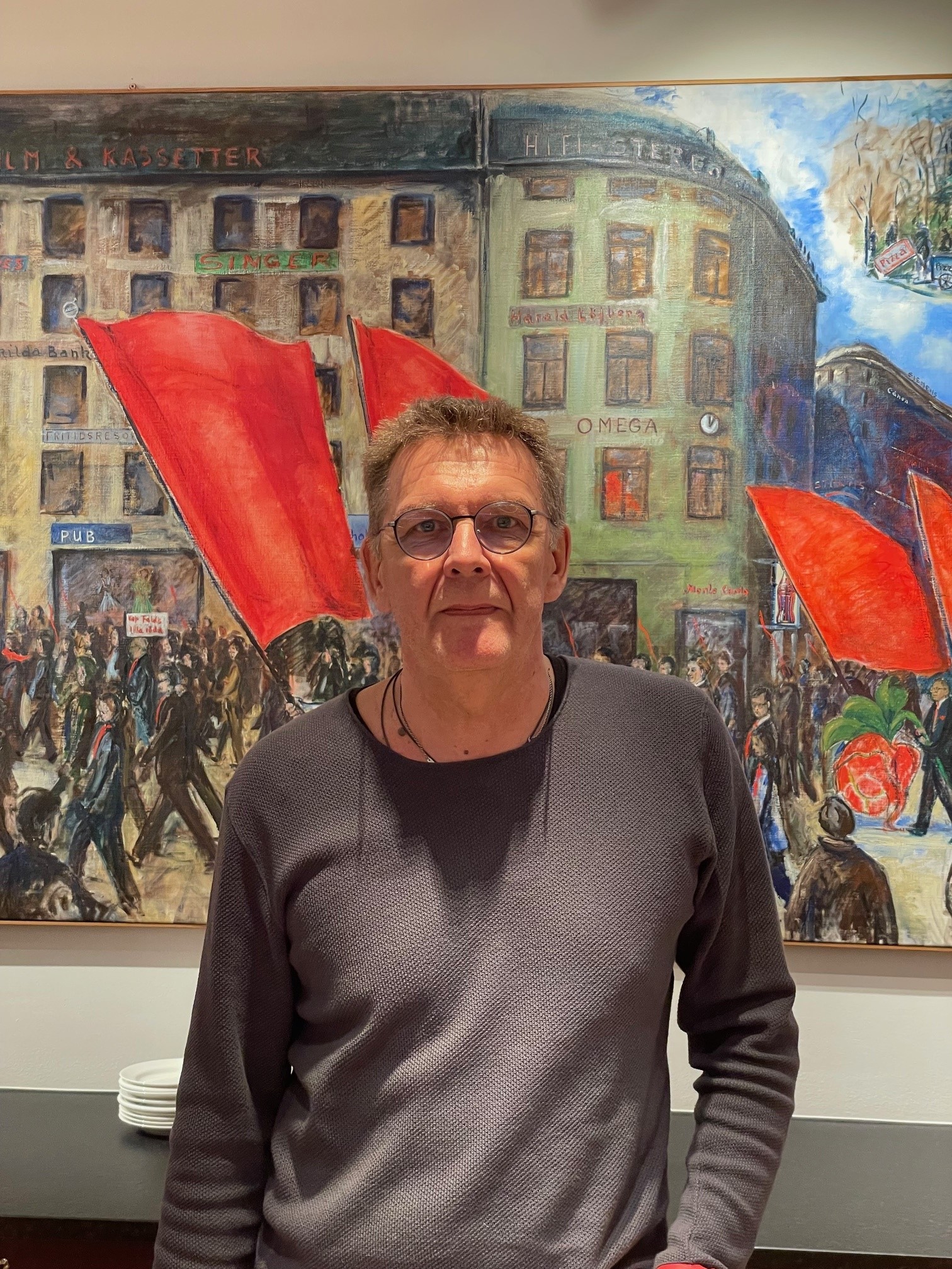 En man med glasögon och grå skjorta står framför en muralmålning som visar en livlig stadsmiljö med röda flaggor.