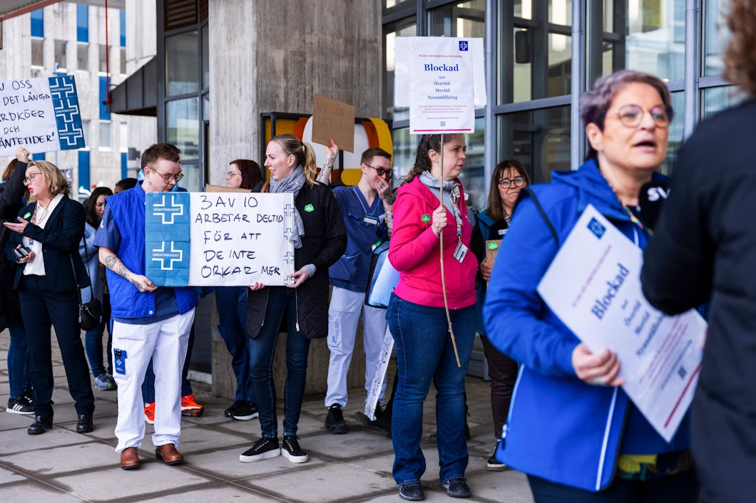 Folksamling utanför en byggnad, personer håller upp skyltar med text på svenska. Några bär labbrockar, andra vanliga kläder. En tydlig skylt beskriver arbetsvillkor.