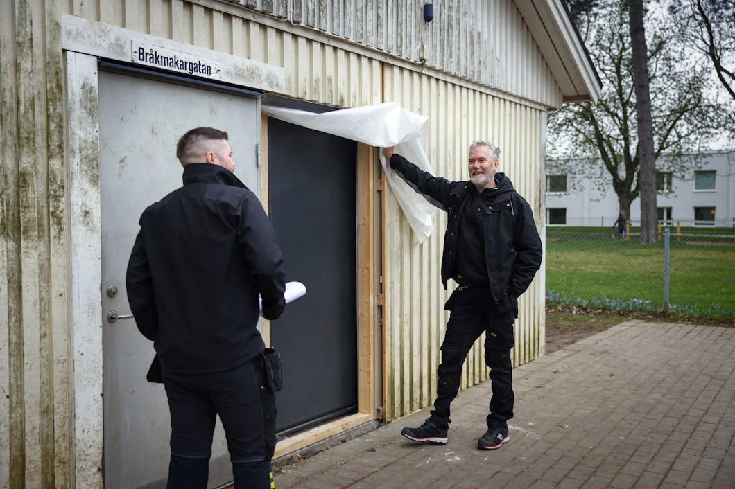 Rikard Jönsson visar skyddsrumsdörren bredvid den vanliga ingången, över vilken det sitter en skylt med texten "Bråkmakargatan". Träd och bostadshus syns i bakgrunden.