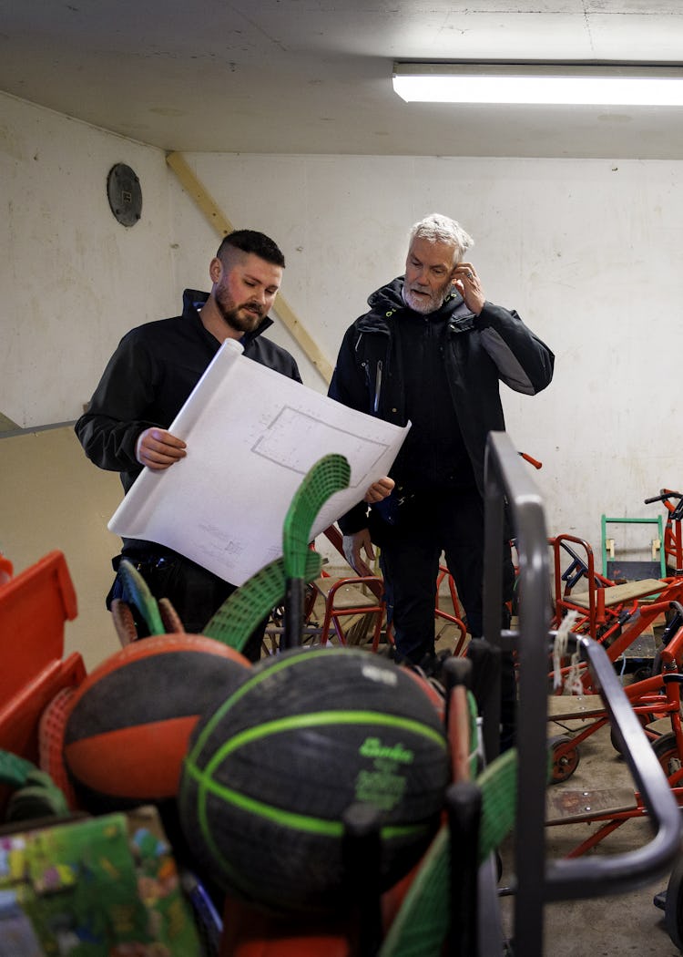 Johan Söderberg och Rickard Jönsson studerar en ritning i ett stökigt skyddsrum med utspridda trehjulingar.