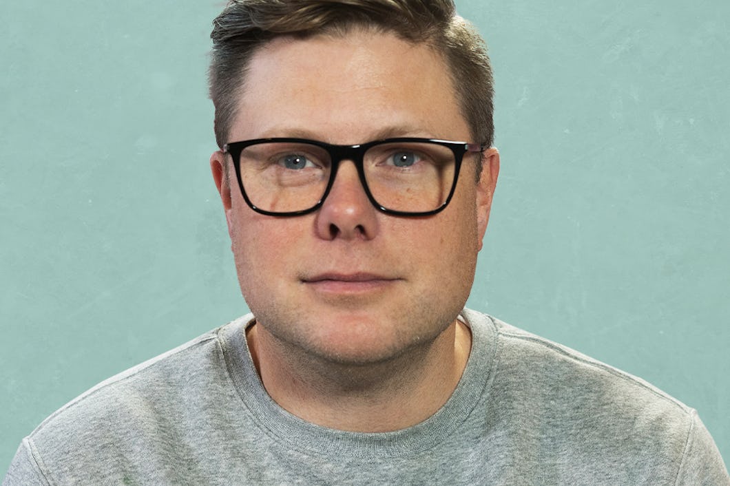 En person med kort hår och glasögon står vänd mot kameran. De har på sig en grå sweatshirt med delvis synlig text. Bakgrunden är ljusblå.