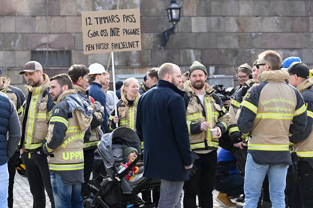 En grupp brandmän och personer är samlade utomhus, några håller en skylt som lyder "12 timmars pass gör oss inte piggare men försörjer familjerna.