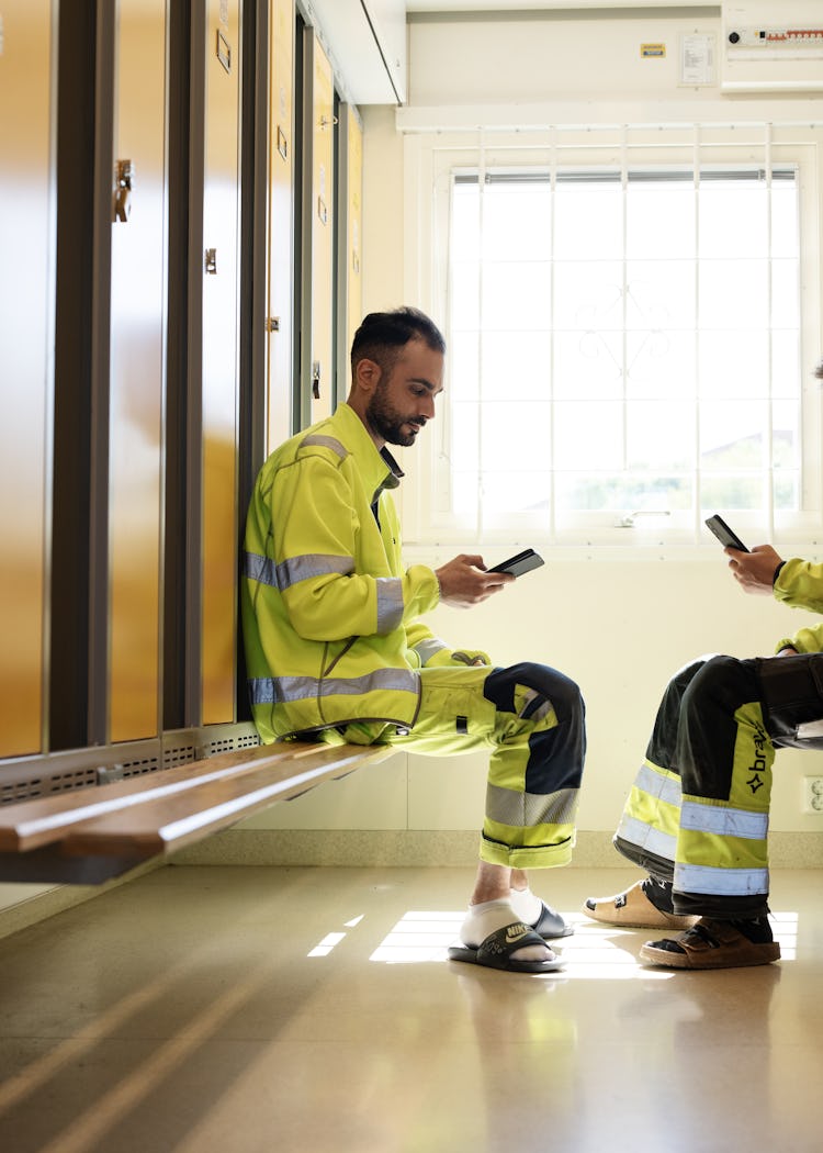 Två arbetare i kläder med hög synlighet sitter på motsatta bänkar i ett omklädningsrum och tittar på sina telefoner.
