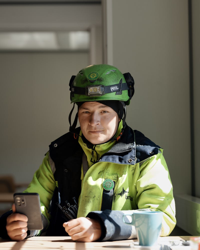 En person med en grön skyddshjälm och gul jacka sitter vid ett bord och håller en smartphone.