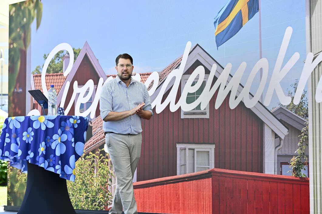 En man står på scenen och applåderar. I bakgrunden finns svensk text och en husbild. En hylla med en blomsterprydd duk och en vattenflaska står bredvid honom.