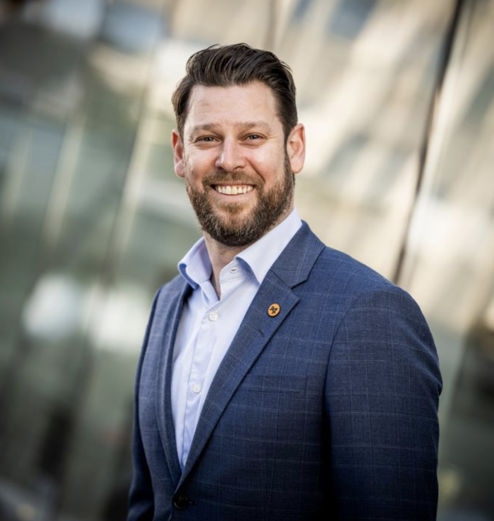 Jonas Jegers med skägg och kort hår, klädd i en ljusblå skjorta och en mörkblå kavaj, står framför en suddig reflekterande bakgrund.