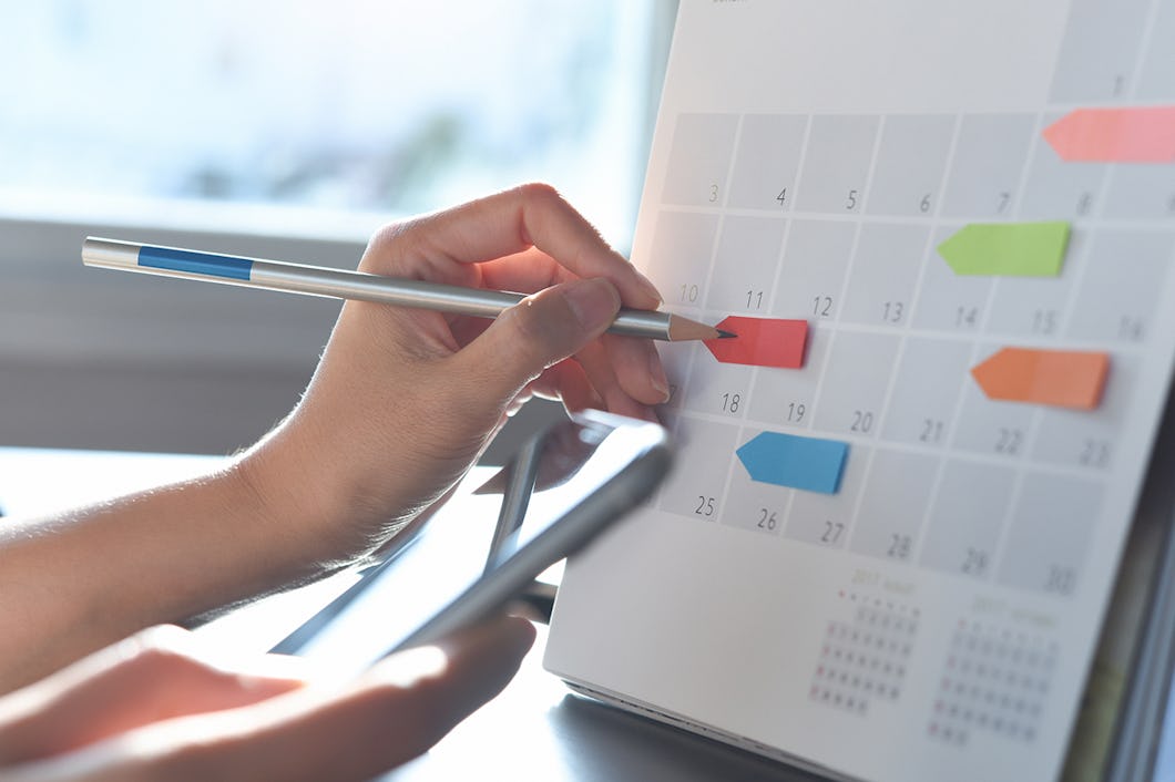 En person använder en penna för att markera ett datum på en kalender och håller samtidigt i en smartphone. Färgglada flikar är fästa vid olika datum på kalendern.