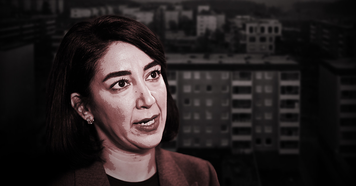 En kvinna med mörkt axellångt hår talar framför en stadsmiljö med flerfamiljshus.